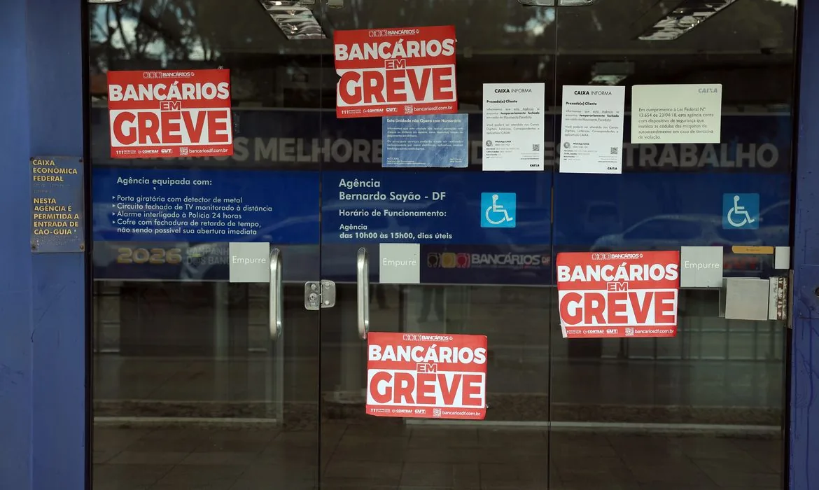 Cartazes vermelhos de bancários anunciam a greve da Caixa na porta de vidro de agência fechada no Distrito Federal