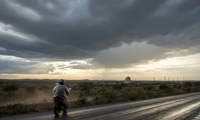 da imagem de destaque Ilustração gerada por inteligência artificial: motociclista de capacete e capa clara, com um garupa, entra numa rodovia asfaltada molhada levantando poeira do acostamento de terra. À frente, uma nuvem escura de tempestade cobre o céu, com cortina de chuva à direita e uma faixa de luz alaranjada do sol baixo à esquerda. Nos dois lados, vegetação rasteira e árvores esparsas; ao fundo, uma torre branca e linhas de transmissão.