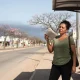 Mulher de camiseta verde se abana com um jornal dobrado em ponto de ônibus de avenida vazia, sob sol forte, entre árvores floridas sem folhas e uma faixa de nuvem escura no horizonte.