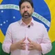 Delegado Sérgio Ribeiro, de barba e camisa rosa clara, fala à câmera diante de uma bandeira do Brasil