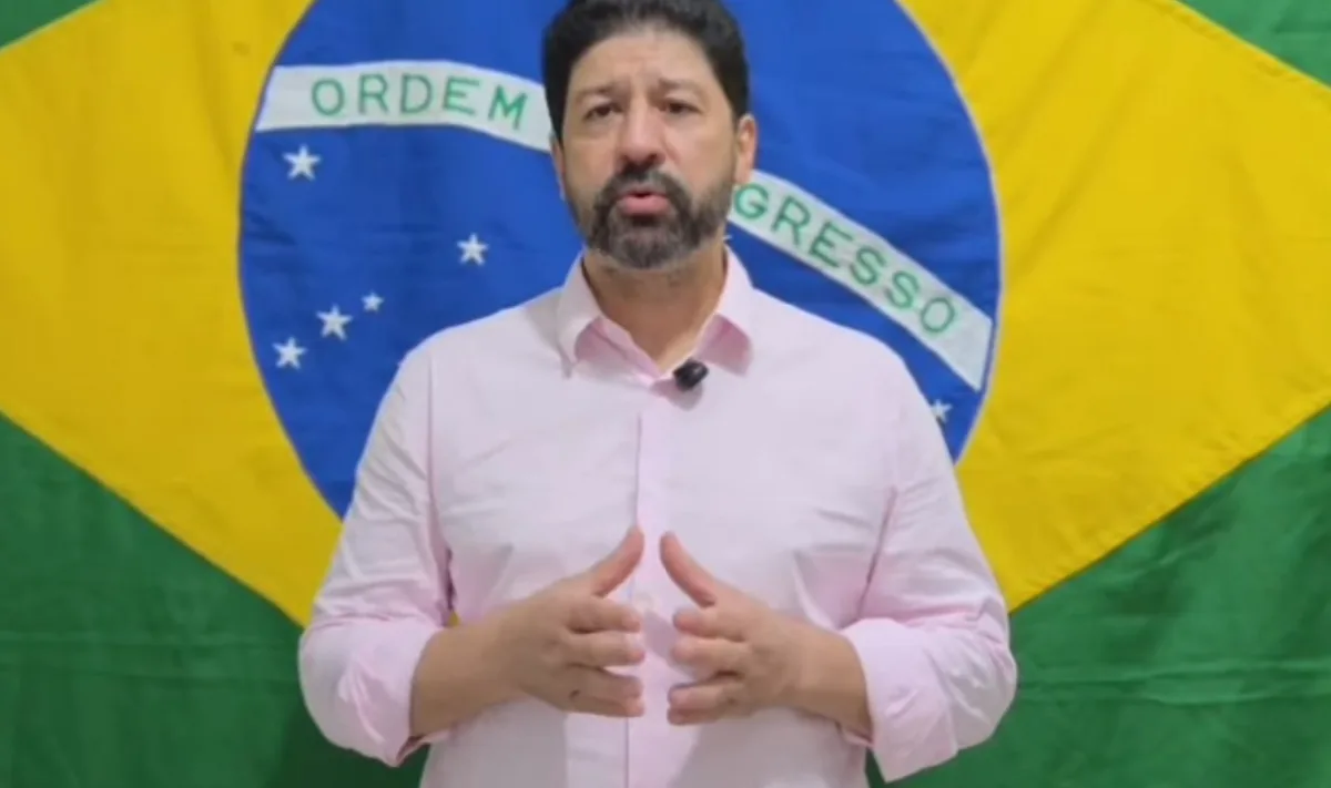 Delegado Sérgio Ribeiro, de barba e camisa rosa clara, fala à câmera diante de uma bandeira do Brasil