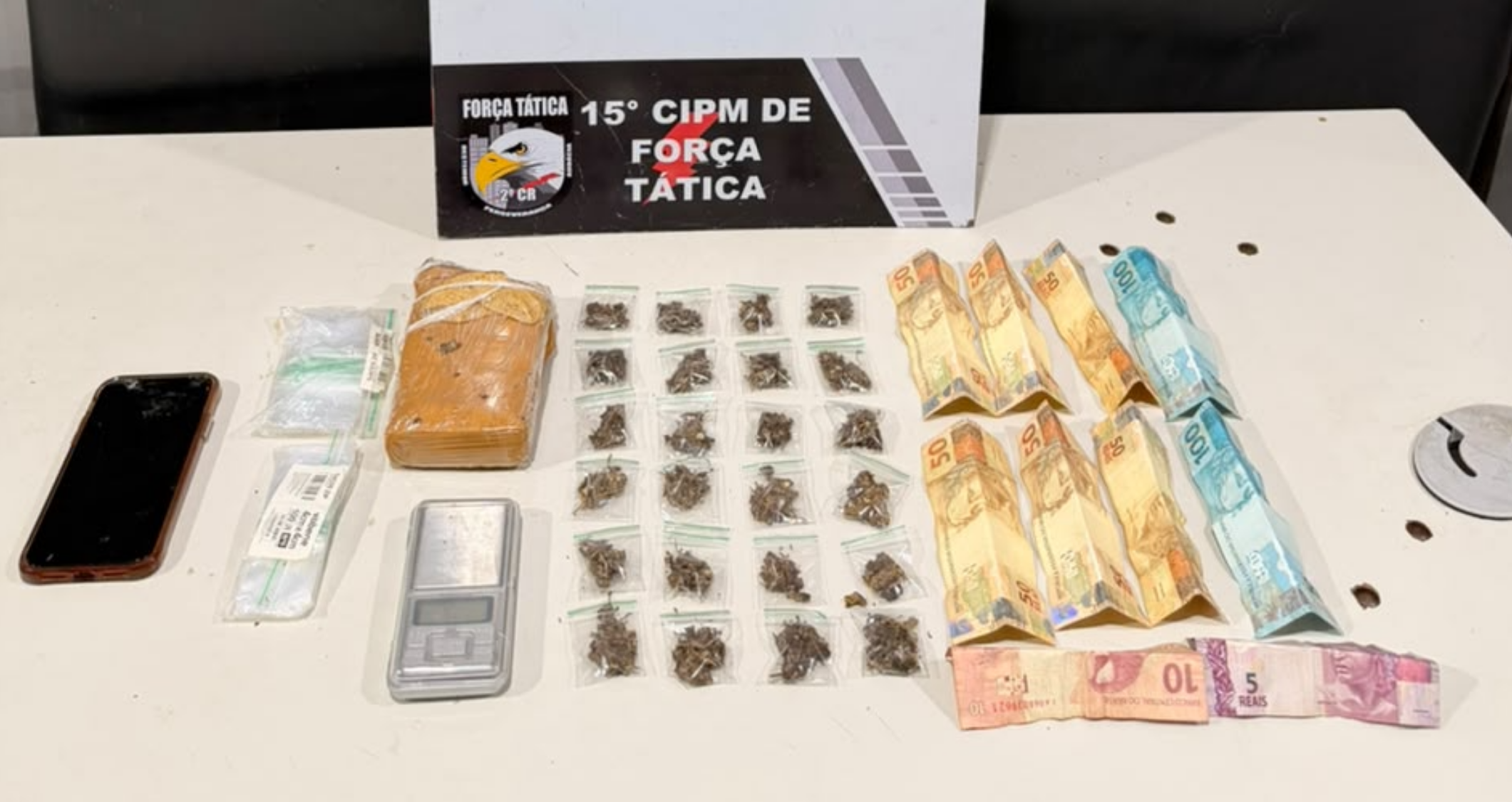 Tablete e porções de maconha, balança de precisão e cédulas de dinheiro sobre uma mesa, diante de placa da 15ª CIPM de Força Tática